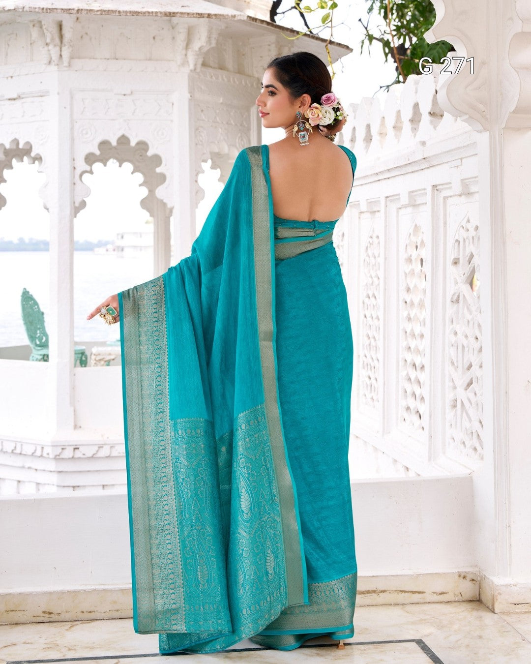 A woman wearing a teal blue-green saree with a gold border, standing in a white architectural courtyard and photographed from the back.