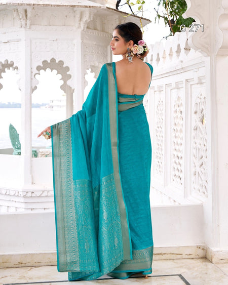 A woman wearing a teal blue-green saree with a gold border, standing in a white architectural courtyard and photographed from the back.