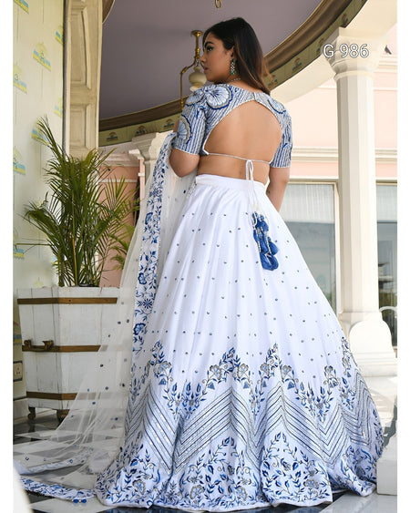 A woman modeling a white and blue printed traditional dress with a flared skirt, short patterned sleeves, and an open back, photographed in an indoor architectural setting with columns and a plant.