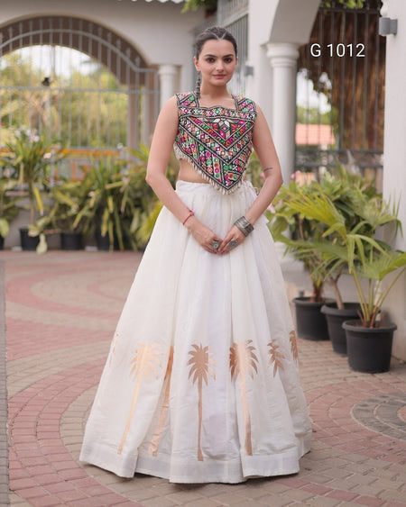 A woman stands outdoors wearing a traditional Indian outfit consisting of a sleeveless white skirt with gold palm tree embroidery and a heavily embroidered multicolor crop top. The background shows a courtyard with potted plants and columns.