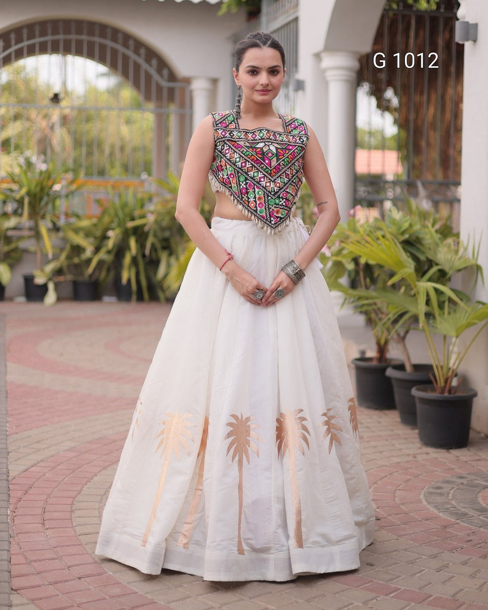 A woman stands outdoors wearing a traditional Indian outfit consisting of a sleeveless white skirt with gold palm tree embroidery and a heavily embroidered multicolor crop top. The background shows a courtyard with potted plants and columns.