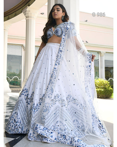 A woman models a white and blue floral lehenga choli set with a matching sheer dupatta in an outdoor architectural setting with white pillars.