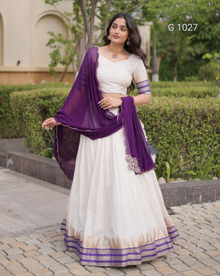 A woman modeling a traditional Indian outfit consisting of a white lehenga skirt, a white blouse with purple sleeve bands, and a draped purple dupatta, set against a garden background. The text 'G 1027' is visible in the top right corner.