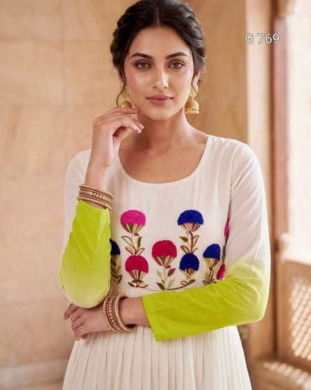 Woman wearing a white outfit with colorful floral embroidery and green sleeves against a beige background.