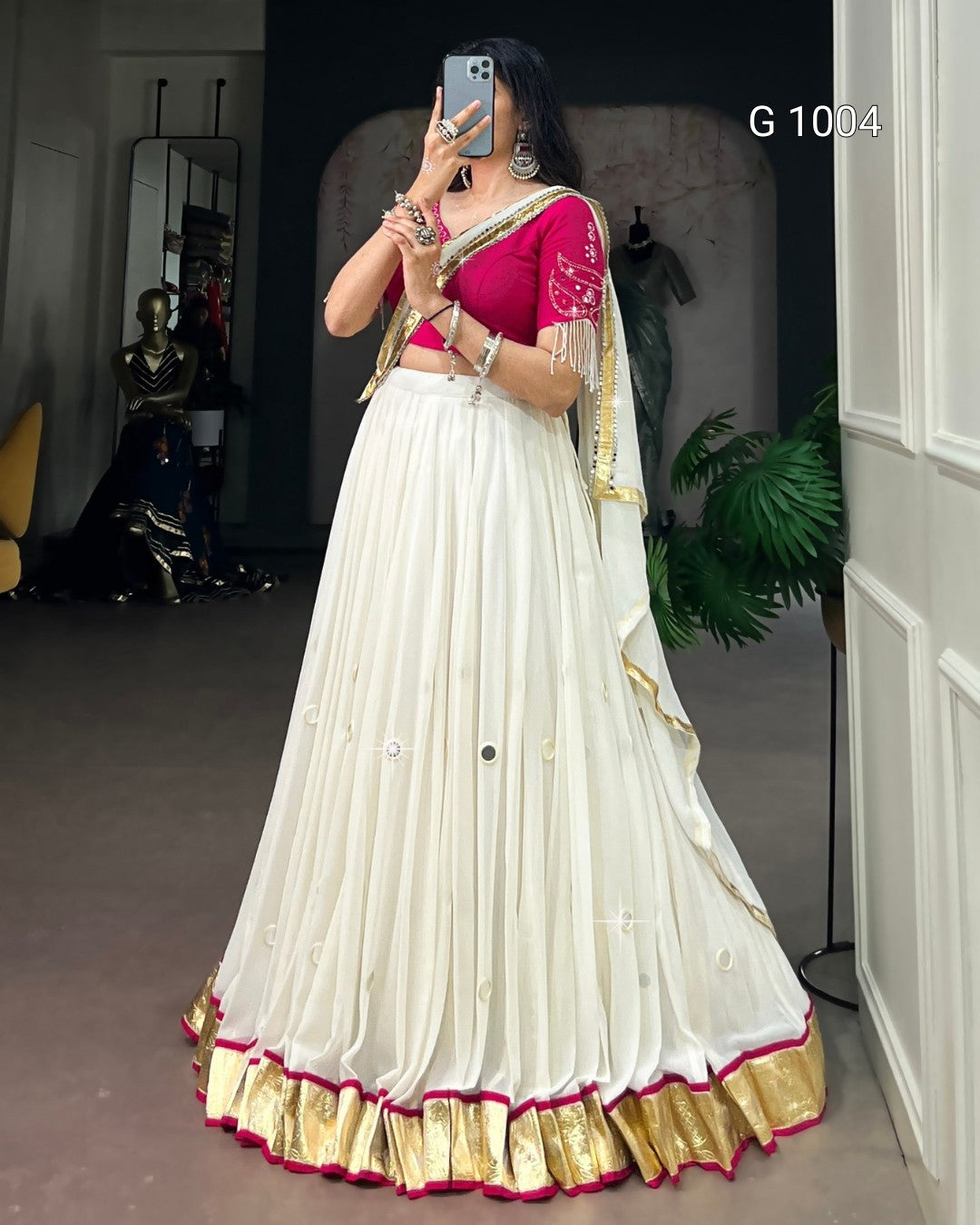 A woman takes a mirror selfie wearing a traditional Indian outfit consisting of a white flared lehenga skirt with gold zari borders, a bright pink blouse with gold embroidery, and a white dupatta with gold detailing. The setting appears to be a boutique interior.