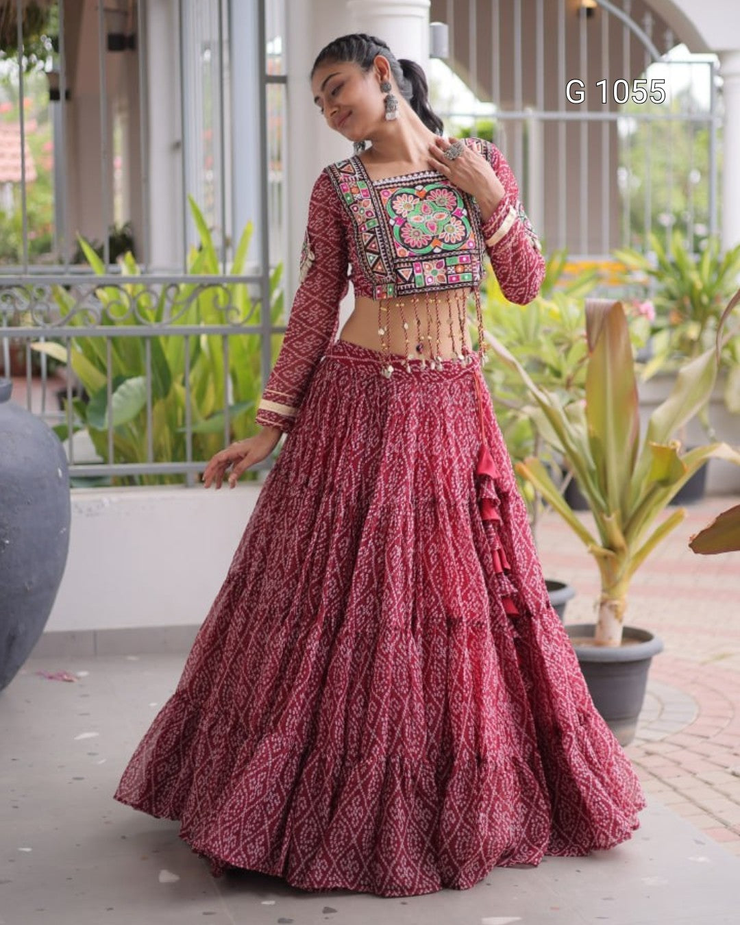 A woman modeling a traditional Indian lehenga choli outfit in a dusty rose pink color with intricate multicolor floral embroidery on the bodice. The outfit features a long flared skirt (lehenga) and a long-sleeved embroidered top (choli) with a small waist cutout and decorative hanging tassels.
