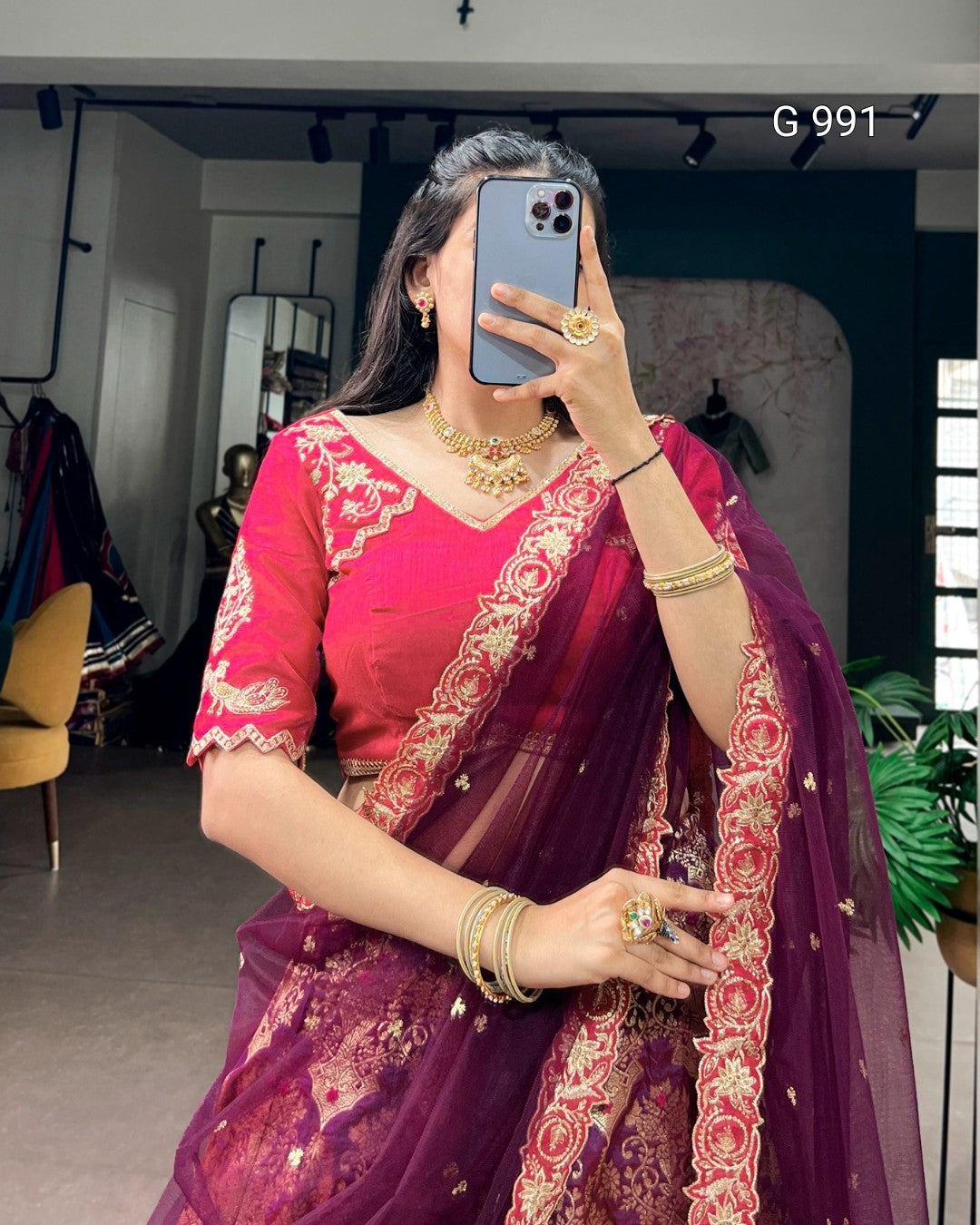 A woman takes a mirror selfie wearing a traditional Indian saree with a bright pink embroidered blouse and a deep purple saree with red floral border embroidery. She is wearing gold jewelry and holding a blue smartphone.
