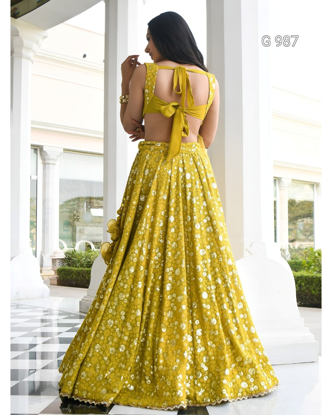 A woman wearing a bright yellow lehenga choli with white floral embroidery, shown from the back in a white architectural corridor. The text 'G 987' is visible in the top right corner.