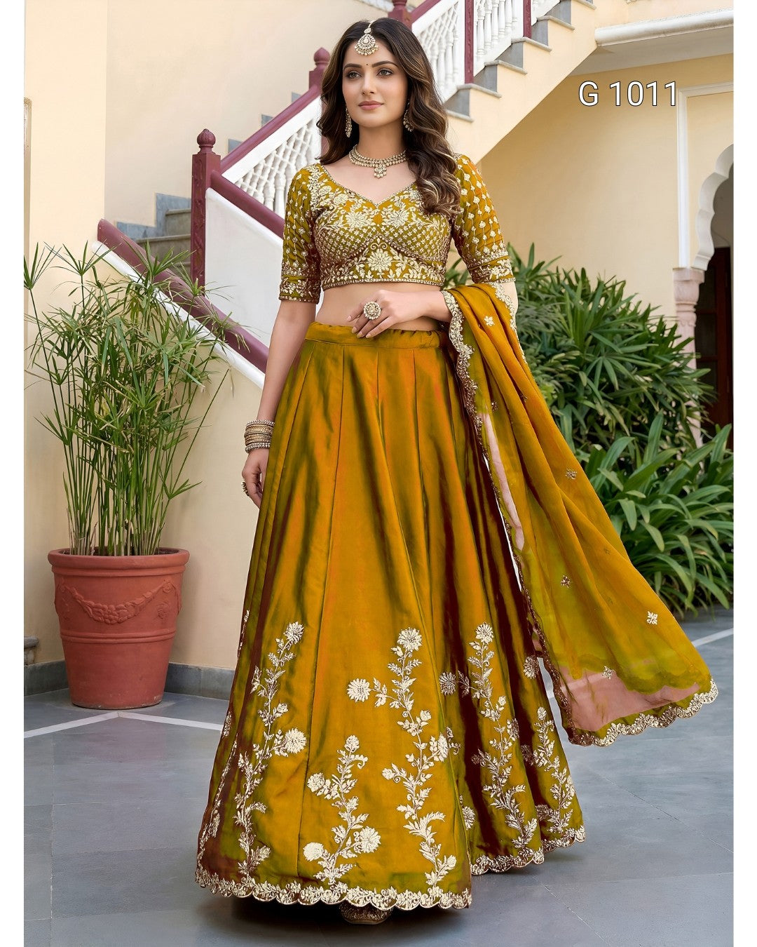 A woman models a traditional Indian lehenga choli suit in mustard yellow with intricate gold and white floral embroidery. The set includes a heavily embellished choli, a full flared skirt, and a matching dupatta draped over her shoulder.