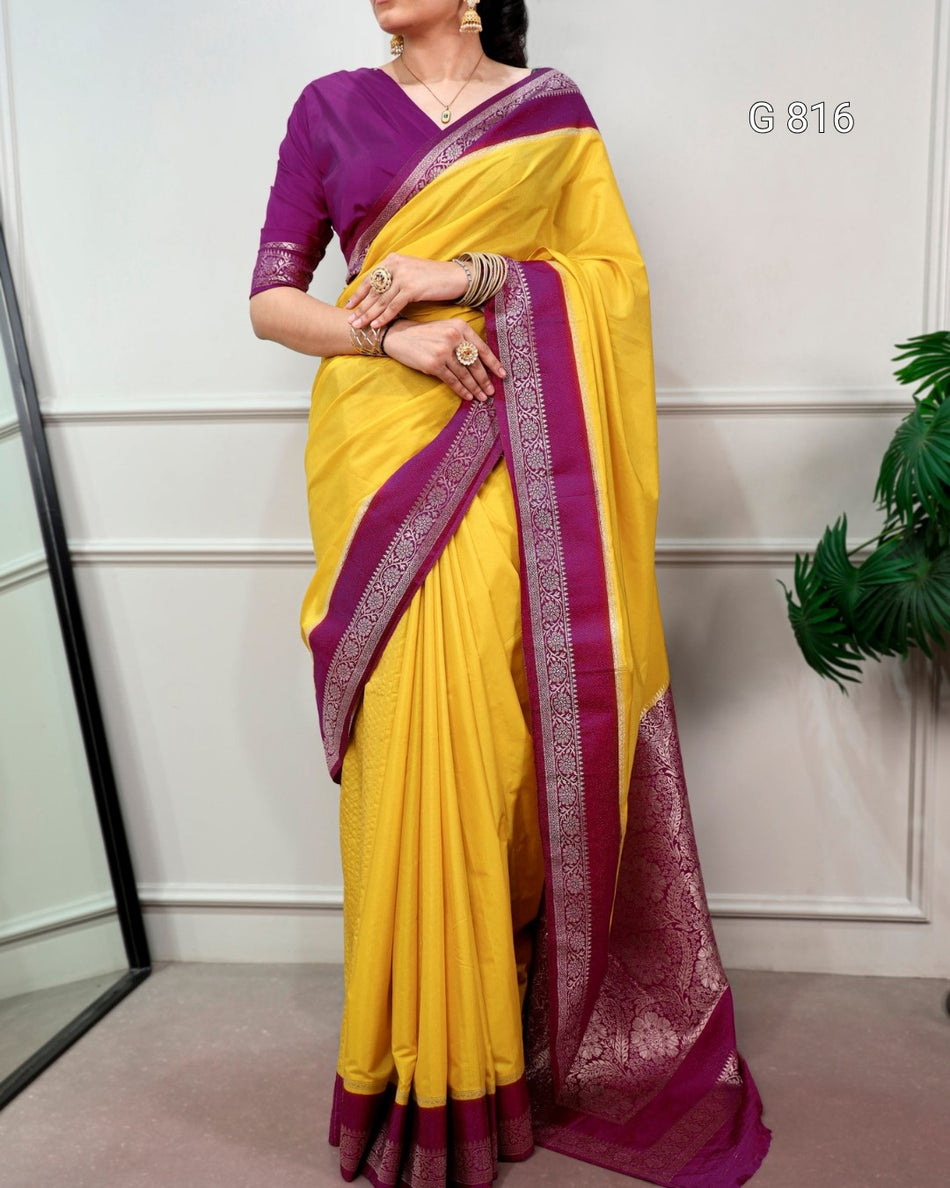 Woman wearing a yellow saree with purple border against a white wall.