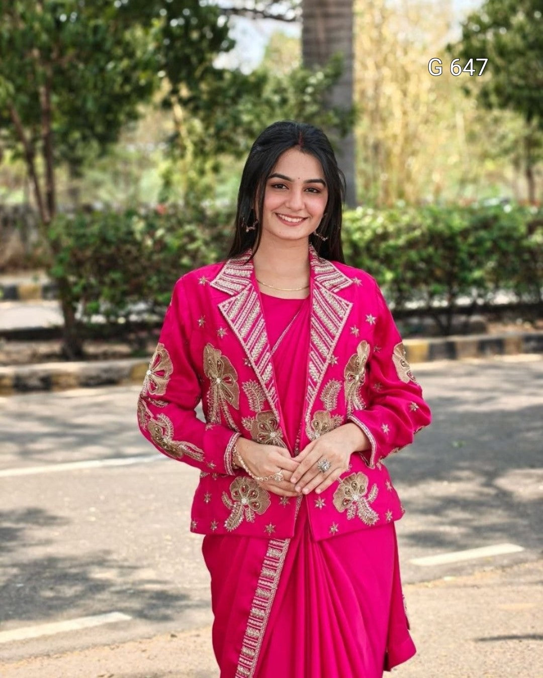 A woman wearing a bright pink traditional outfit with ornate gold embroidery, standing outdoors on a paved path with trees and shrubs in the background.