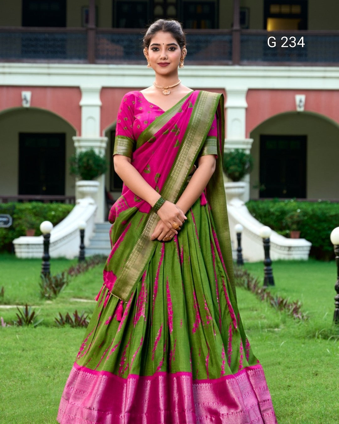Woman in a green and pink saree standing in a garden with a building in the background