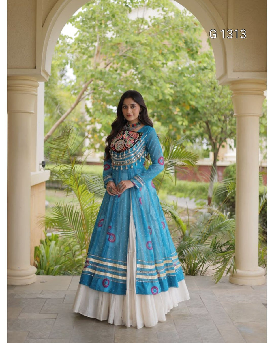 A woman modeling a traditional Indian outfit consisting of a turquoise blue anarkali-style dress with intricate embroidery and a white flared skirt, standing in a courtyard framed by arches.