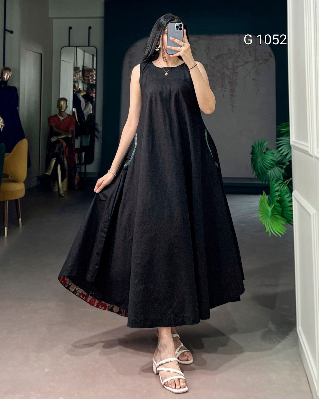A woman takes a mirror selfie in a boutique wearing a sleeveless black A-line maxi dress with a patterned hem and white strappy sandals. The boutique interior has a gray floor, a dark blue wall, and mannequins in the background.