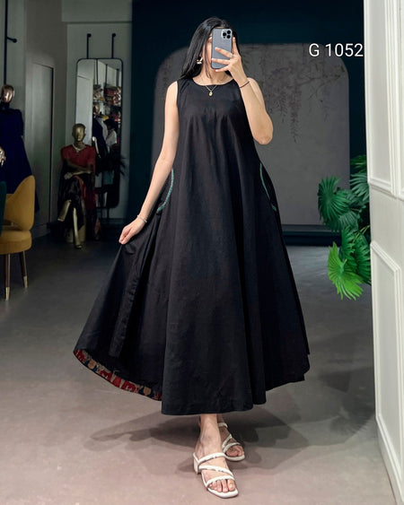 A woman takes a mirror selfie in a boutique wearing a sleeveless black A-line maxi dress with a patterned hem and white strappy sandals. The boutique interior has a gray floor, a dark blue wall, and mannequins in the background.
