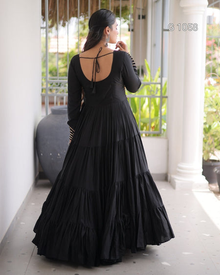 A woman wearing a long black dress with a tiered skirt and strappy open back, standing on a covered porch with columns and greenery in the background.