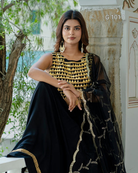A woman wearing a black traditional outfit with a heavily embellished gold embroidered blouse and a sheer black dupatta with gold border detailing, posed outdoors against a stone pillar and greenery.