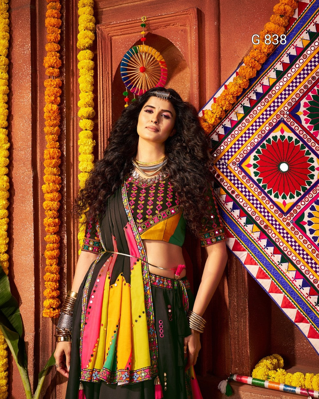 Woman in traditional multicolored outfit standing against a decorated wall with marigold flowers.