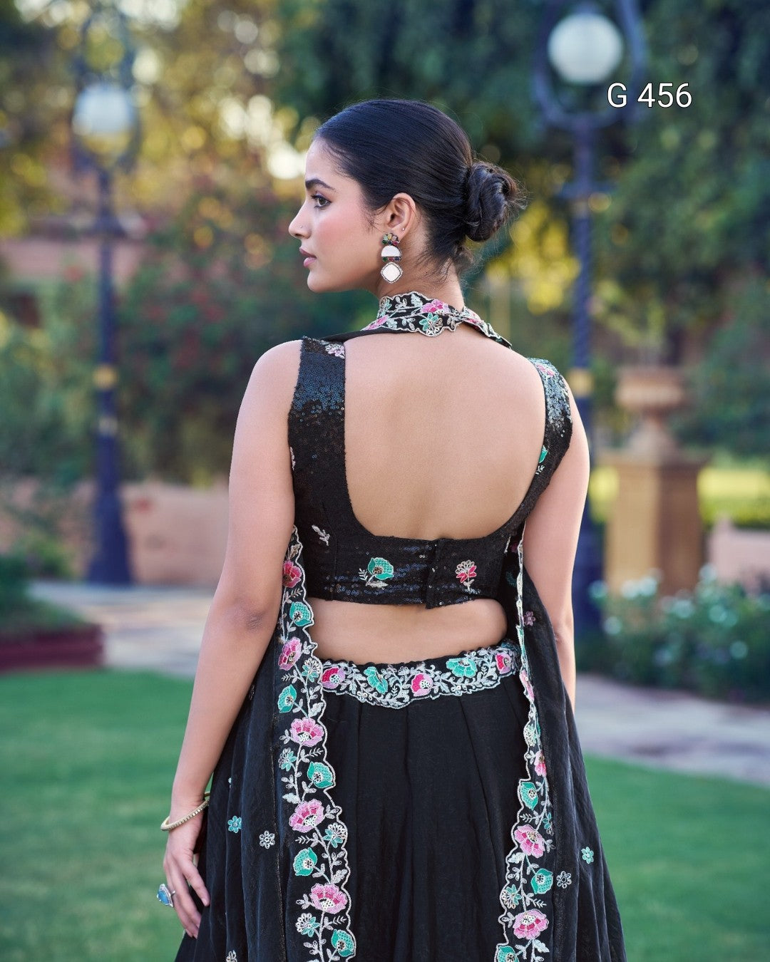 A woman shown from the back wearing a black traditional outfit with colorful floral embroidery, photographed outdoors in a garden setting. The text 'G 456' appears in the top right corner.