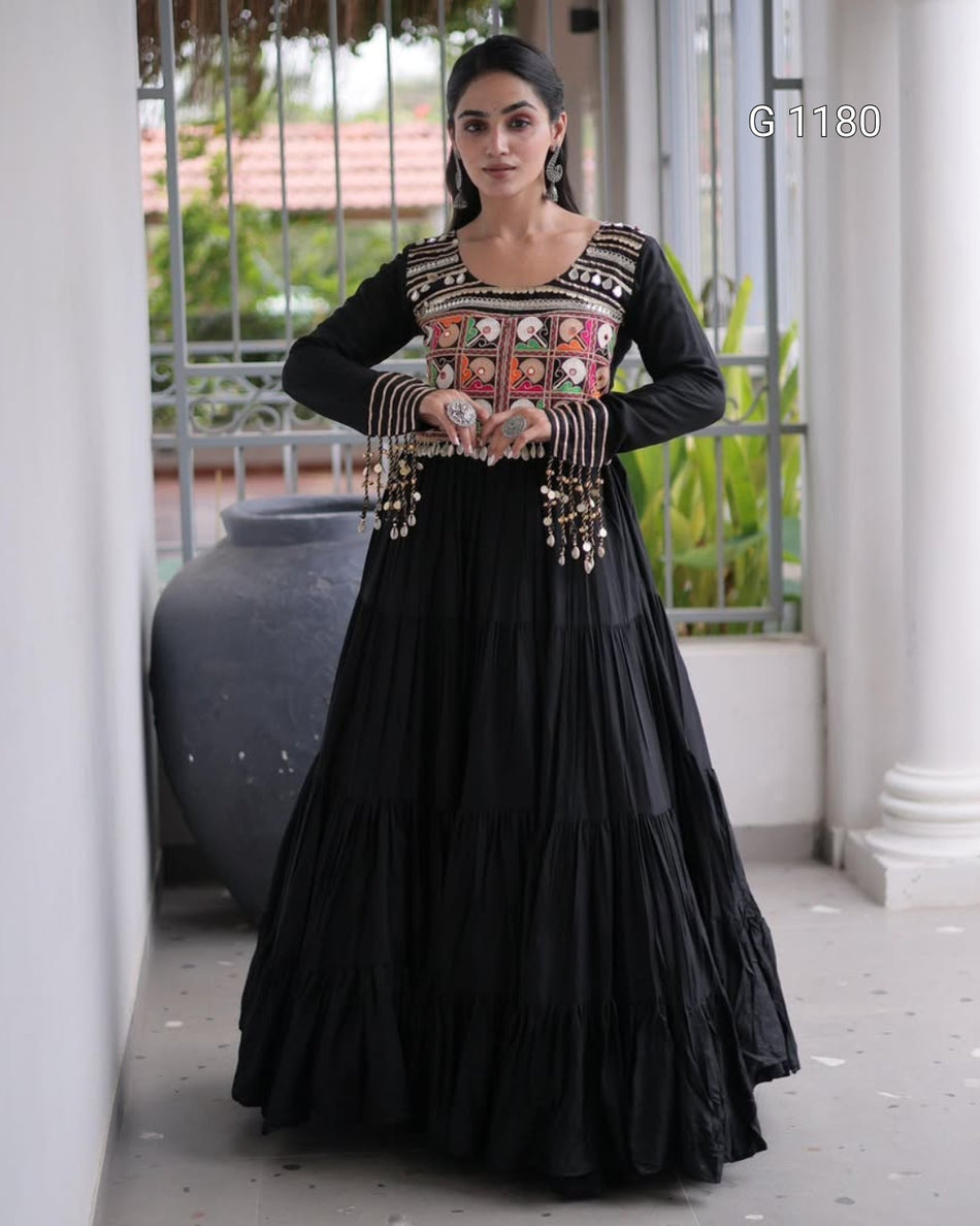 A woman models a black long-sleeve Anarkali gown with a heavily embroidered multicolor bodice and dangling gold-toned embellishments, standing in a covered outdoor walkway.