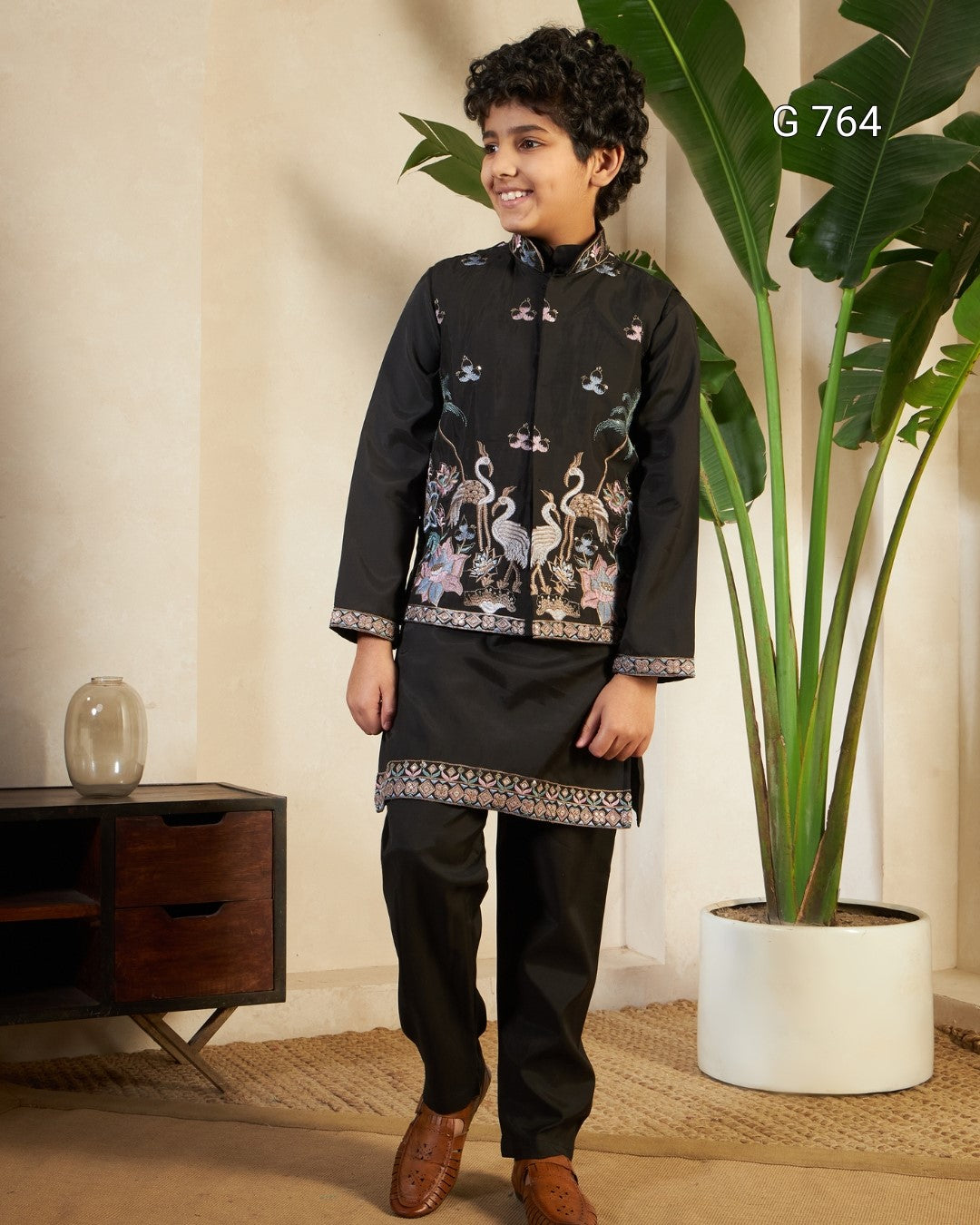 Child wearing a black traditional outfit with intricate patterns in a room with a plant and wooden cabinet.