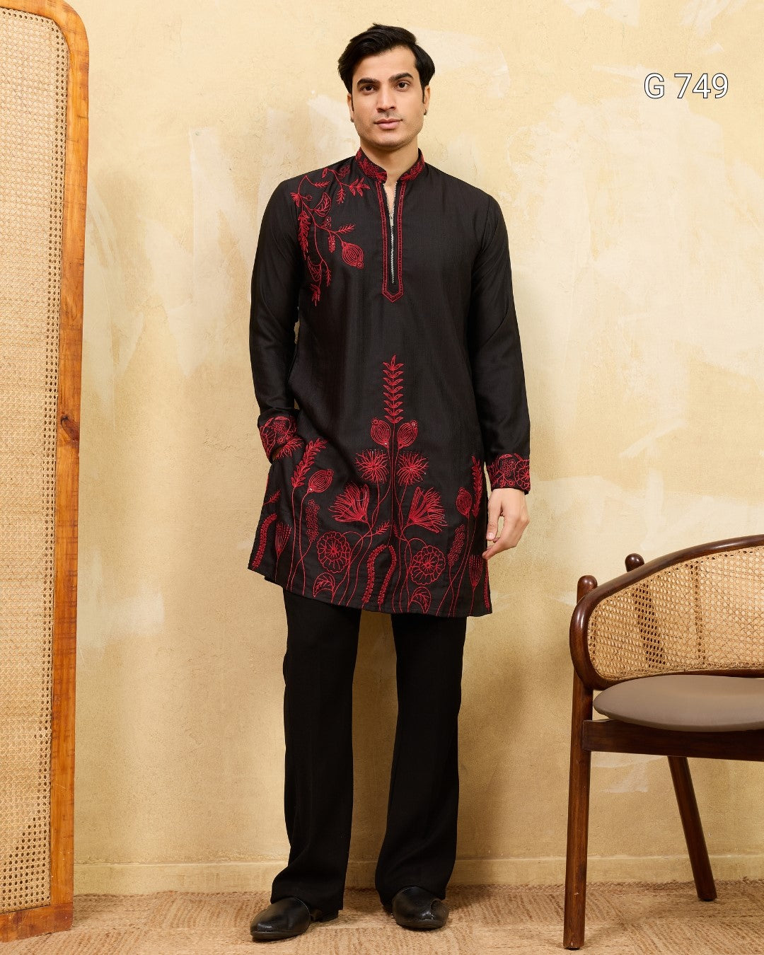 Man wearing a black kurta with red embroidery standing against a beige wall.