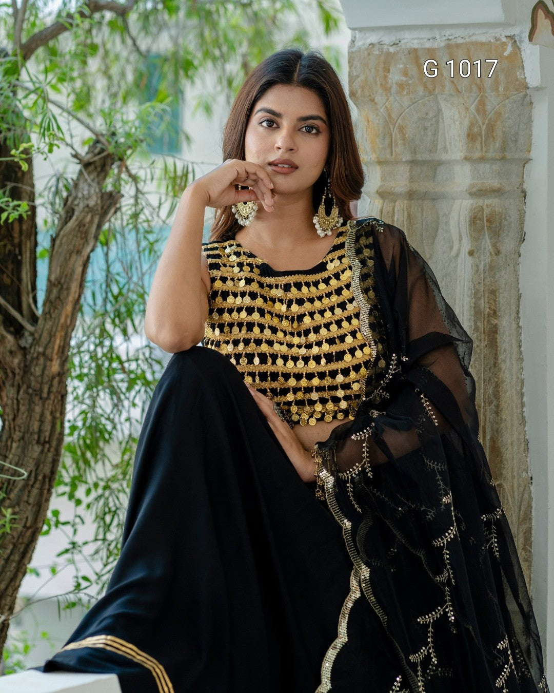 A woman models a black and gold embellished traditional outfit with a sheer black dupatta, seated outdoors against a stone pillar and greenery background. The number G1017 is printed in the top right corner.