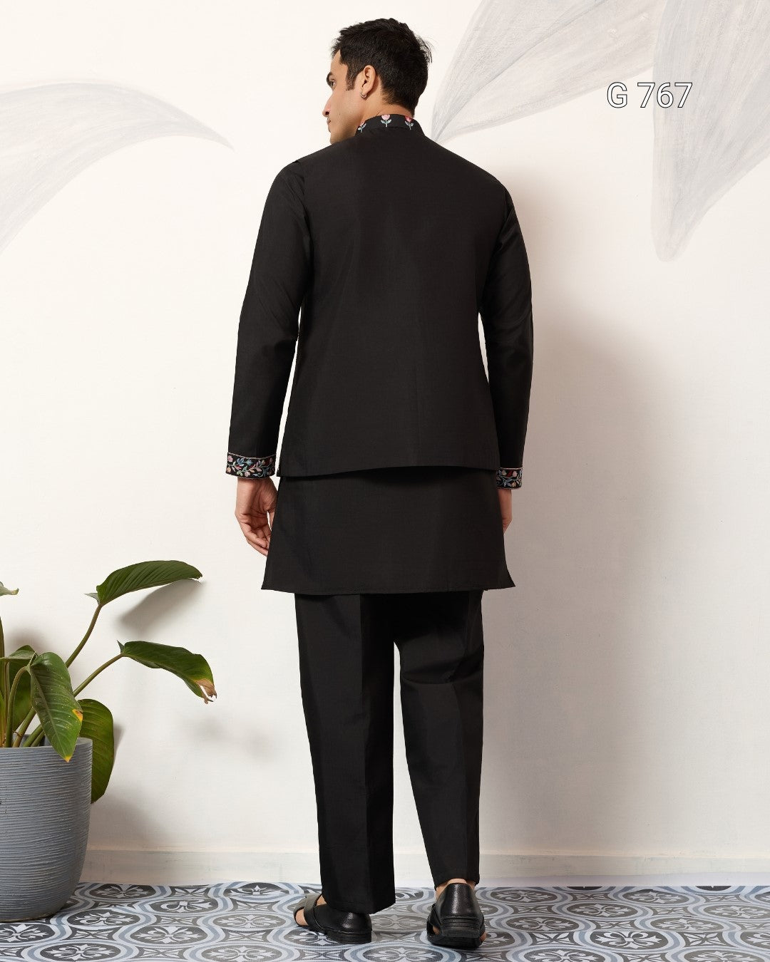 Man wearing a black suit standing in a room with a plant and patterned floor.