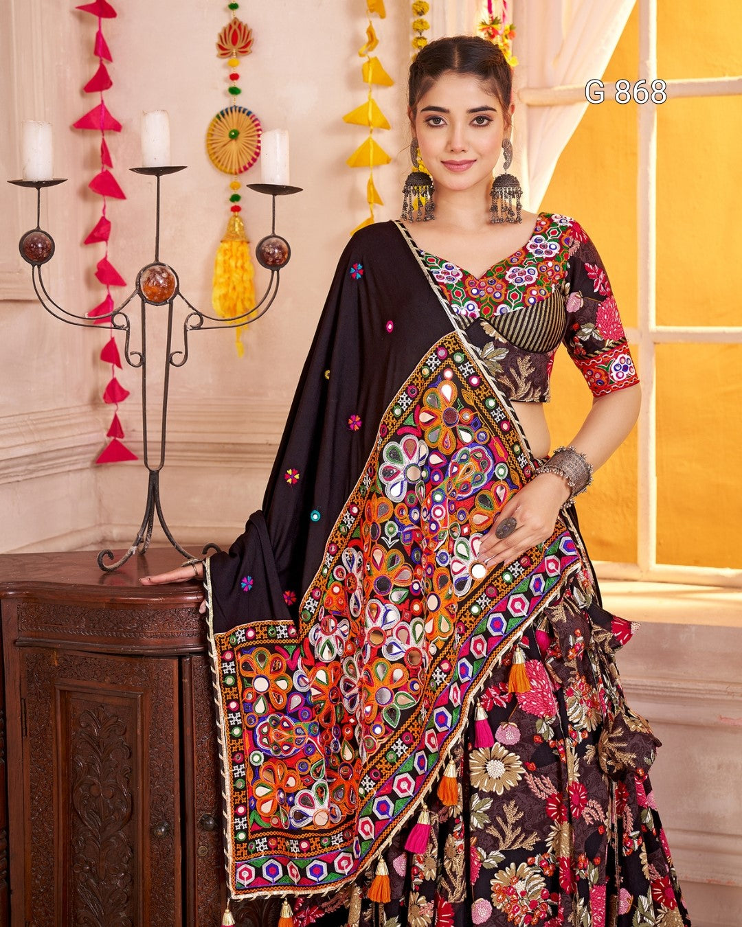 Woman in a colorful traditional outfit with a black dupatta in a decorated room.