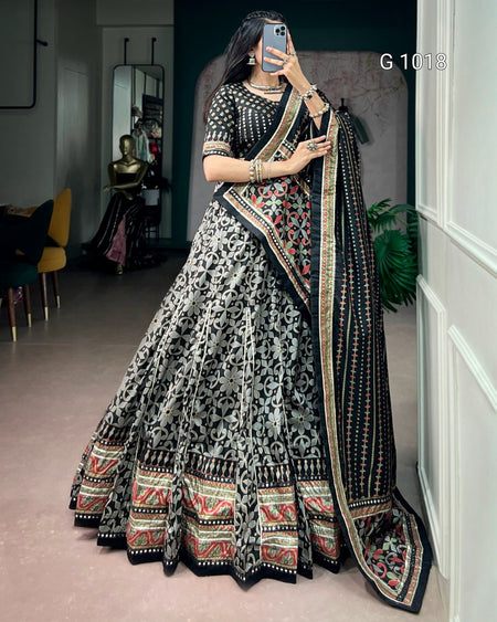 A woman takes a mirror selfie wearing an ornate black and gray printed lehenga choli with a heavily embroidered black dupatta featuring red, green, and gold patterns. The setting is an indoor room with a mirror, chairs, and a plant in the background.