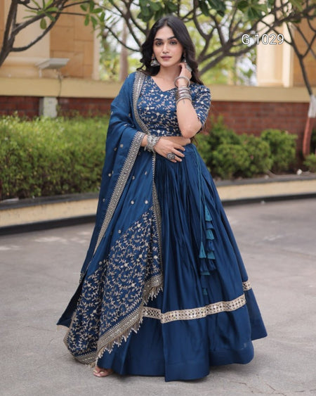 A woman wearing a navy blue traditional Indian dress with silver embroidery, a matching embroidered dupatta, and a flared skirt, standing outdoors in front of a building and greenery.