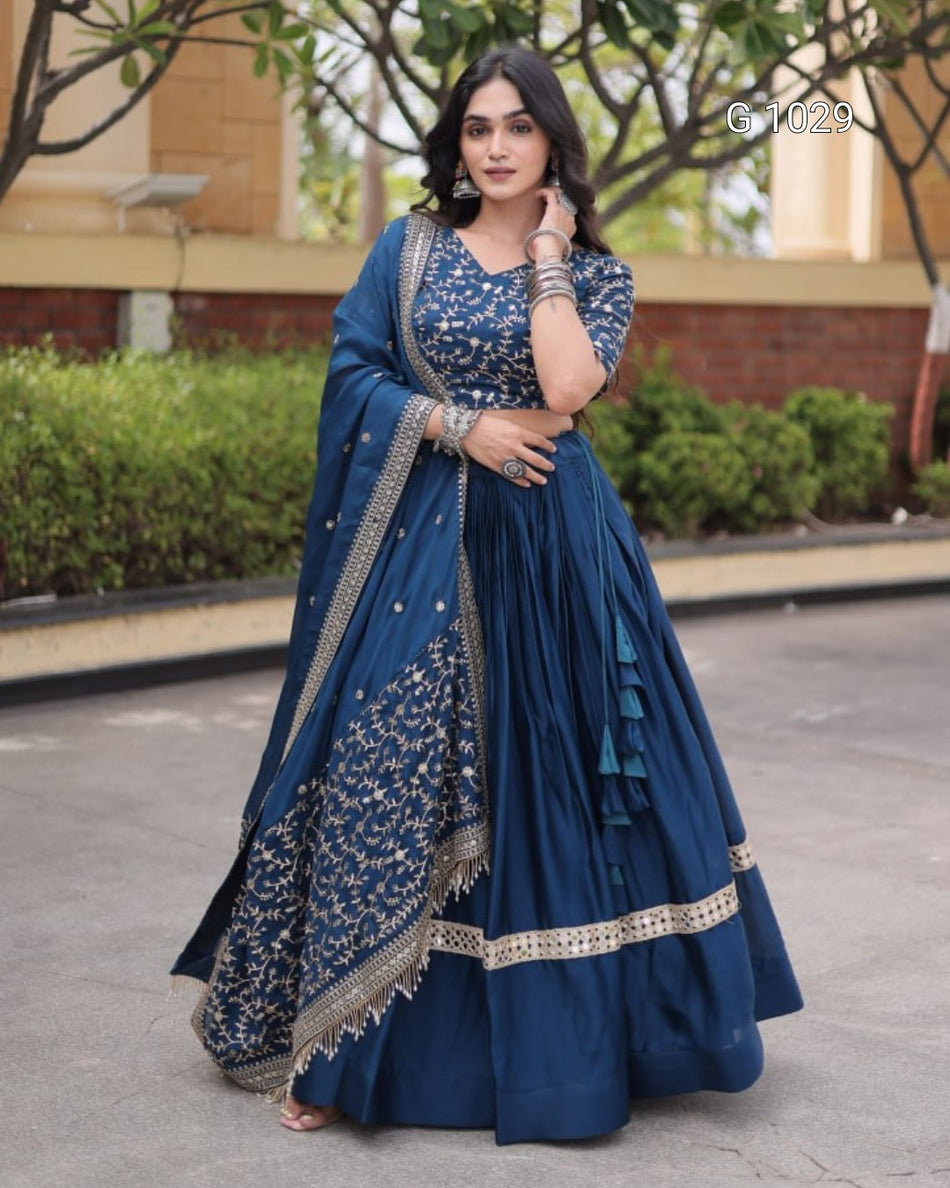 A woman wearing a navy blue traditional Indian dress with silver embroidery, a matching embroidered dupatta, and a flared skirt, standing outdoors in front of a building and greenery.