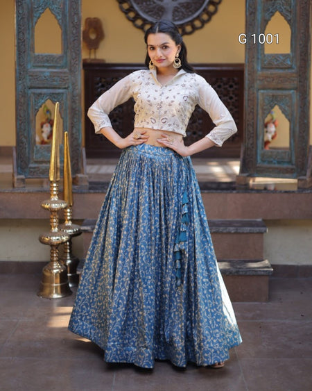 A woman models a traditional Indian outfit consisting of a cream-colored embroidered blouse and a long blue floral printed skirt, standing in front of an ornate architectural background.