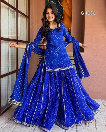 A woman modeling a royal blue traditional Indian outfit with white printed motifs and silver border details, featuring a flared skirt, long sleeves, and a matching dupatta. The image is set against a warm-toned indoor background with a window and wall art, and the text 'G 1030' is visible in the top right.