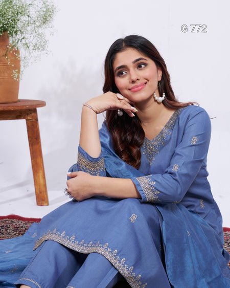 Woman wearing a blue traditional outfit with silver embroidery, sitting on a white background.