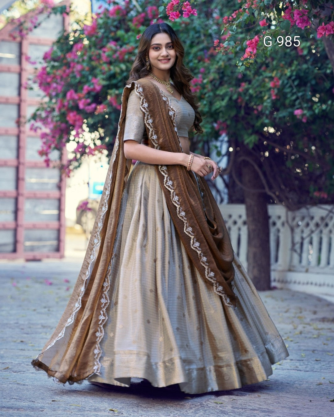 A woman modeling a traditional Indian lehenga choli outfit in a garden setting. The outfit consists of a beige and brown skirt with a matching brown dupatta featuring white embroidery.