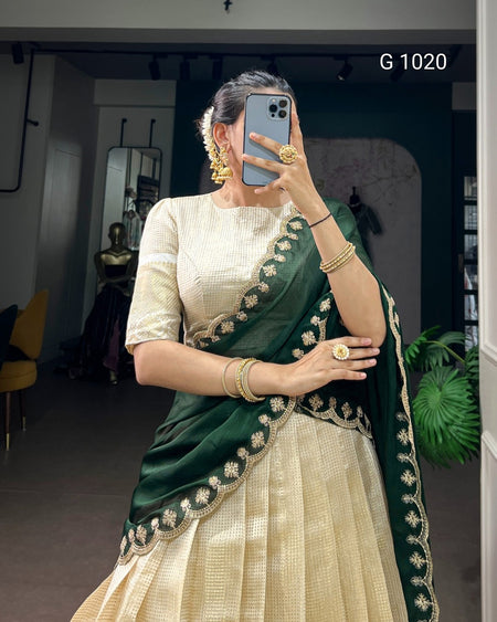 A woman takes a mirror selfie wearing a traditional Indian outfit consisting of a cream-colored textured skirt and a dark green dupatta with gold floral border embroidery. She is wearing gold jewelry, including earrings, rings, and bangles, and is holding a blue smartphone.