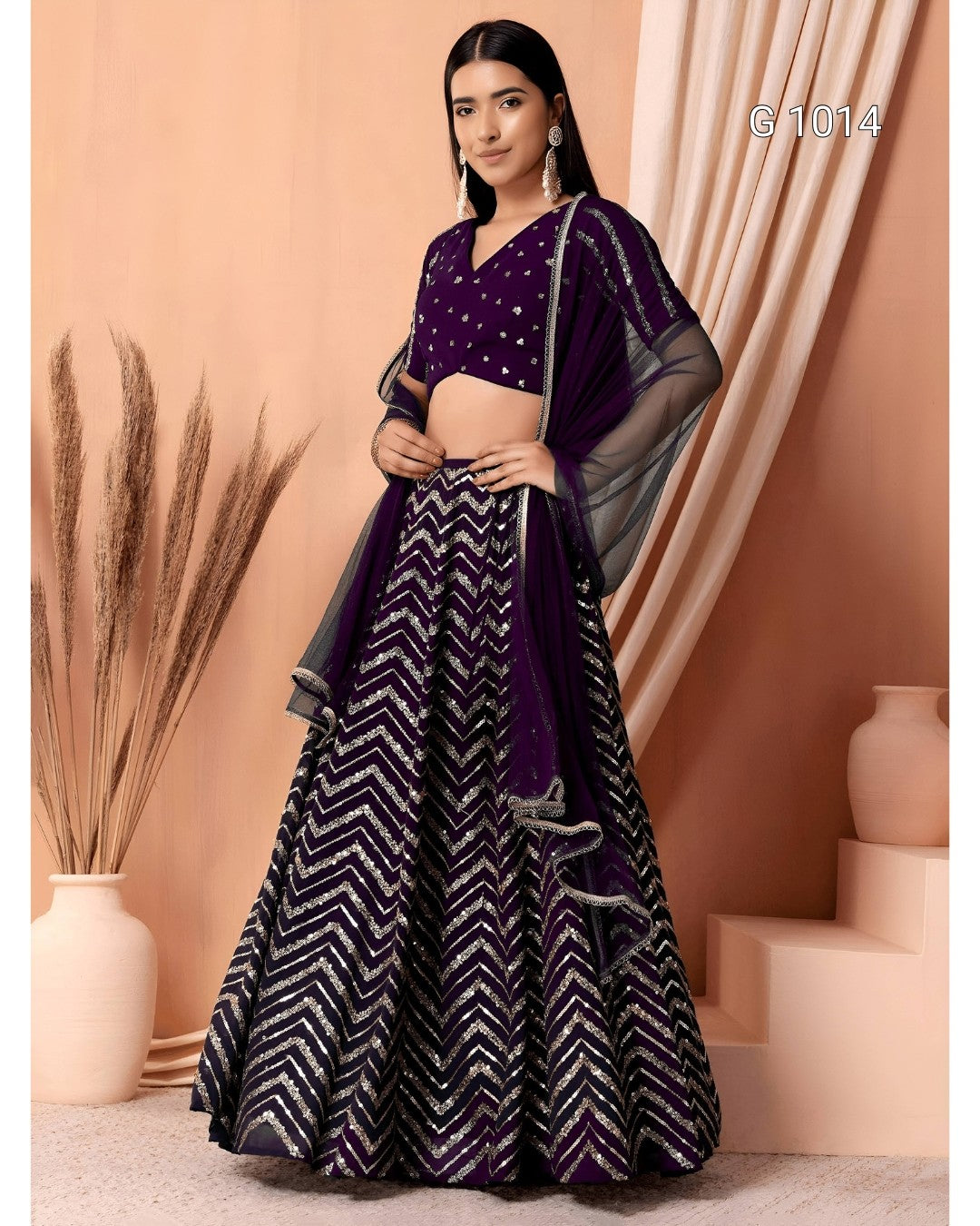 A woman models a traditional Indian outfit consisting of a deep purple embroidered blouse, a matching patterned skirt with silver chevron designs, and sheer dark gray dupatta pieces, set against a warm beige backdrop with decorative vases.