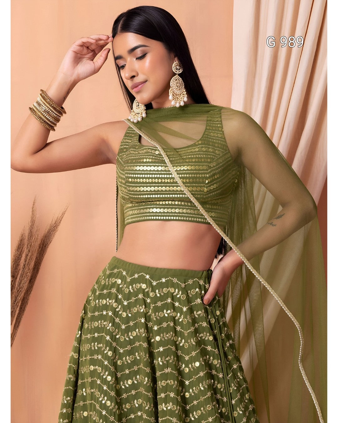 A woman models an olive green ethnic lehenga choli set with gold embroidery, featuring a sheer mesh sleeveless crop top, a flared embroidered skirt, and a matching sheer dupatta draped over her shoulder.