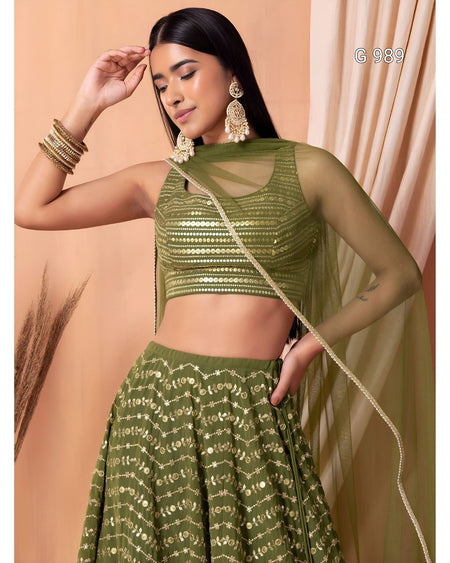 A woman models an olive green ethnic lehenga choli set with gold embroidery, featuring a sheer mesh sleeveless crop top, a flared embroidered skirt, and a matching sheer dupatta draped over her shoulder.