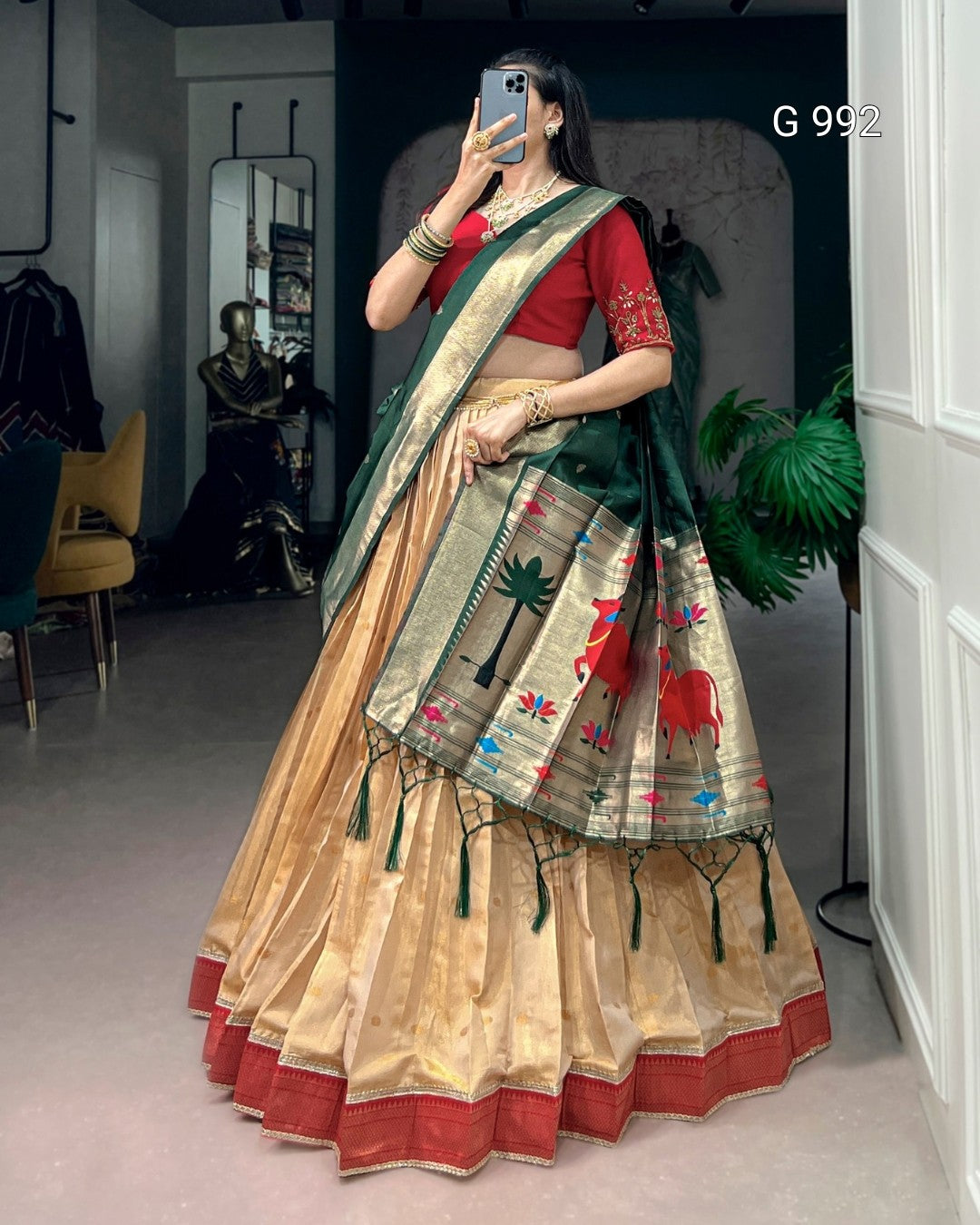 A woman wearing a traditional Indian saree in a mirror selfie inside a boutique. The saree features a beige and gold zari body with red borders, a dark green border, and colorful elephant motifs, paired with a red blouse.