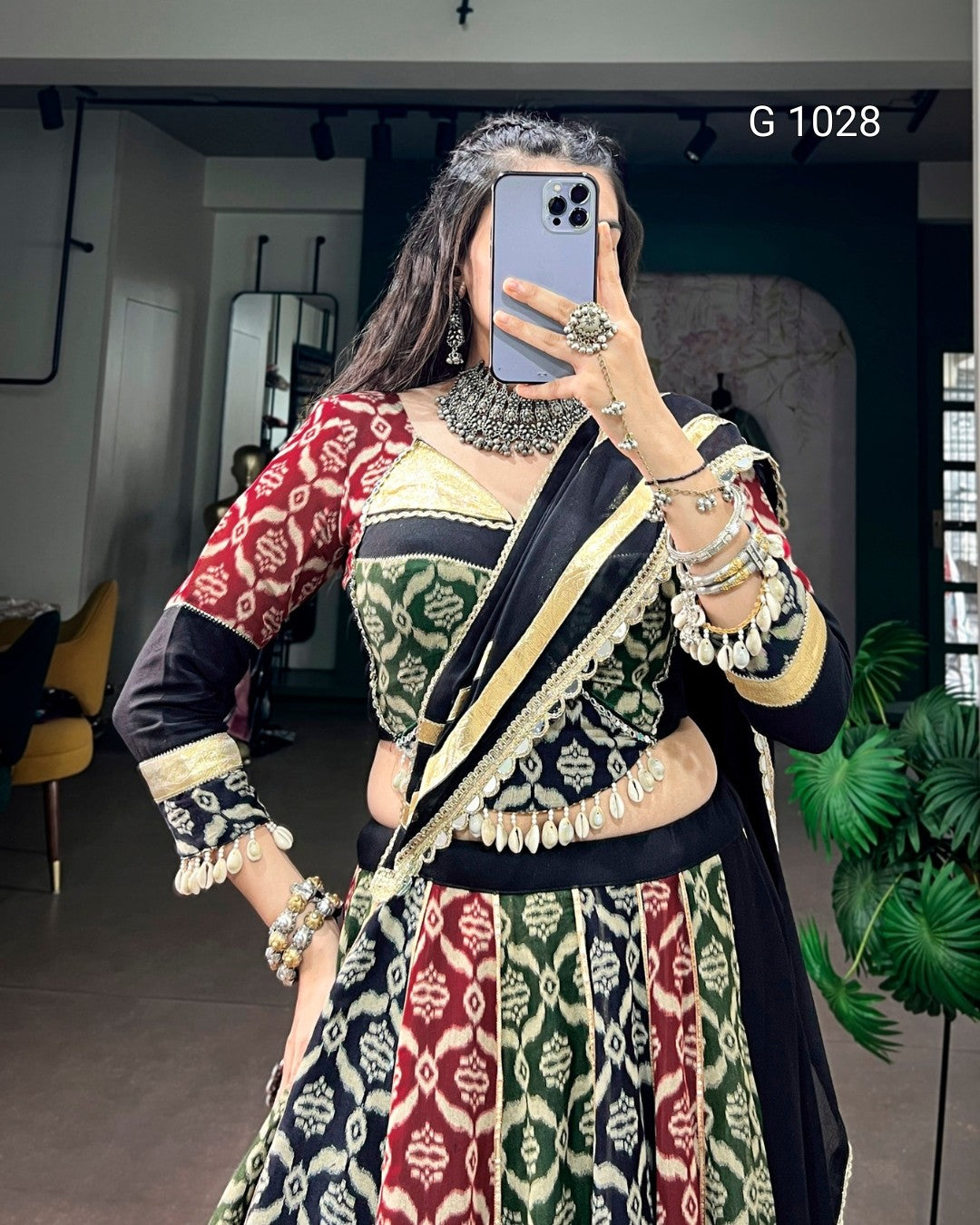 A woman takes a mirror selfie wearing a traditional Indian ethnic outfit with a heavily patterned kurta and matching blouse. The outfit features red, green, black, and cream colors with intricate geometric designs and gold embroidery.