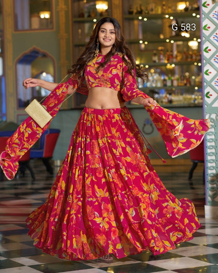 A woman modeling a bright pink and orange floral printed lehenga choli set in an indoor decorative setting. The outfit features a crop top with long bell sleeves and a matching flared skirt.