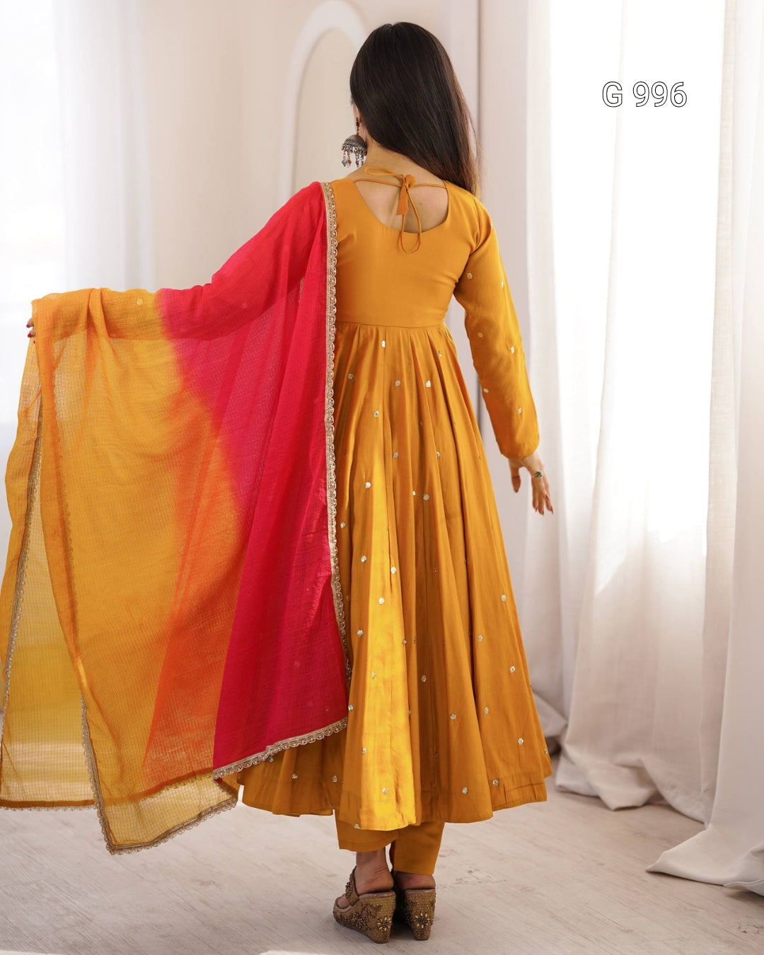 A woman shown from the back wearing a mustard yellow Anarkali gown with small silver embellishments and holding a bright red and yellow dupatta with gold border. She is standing in a brightly lit room with white curtains.