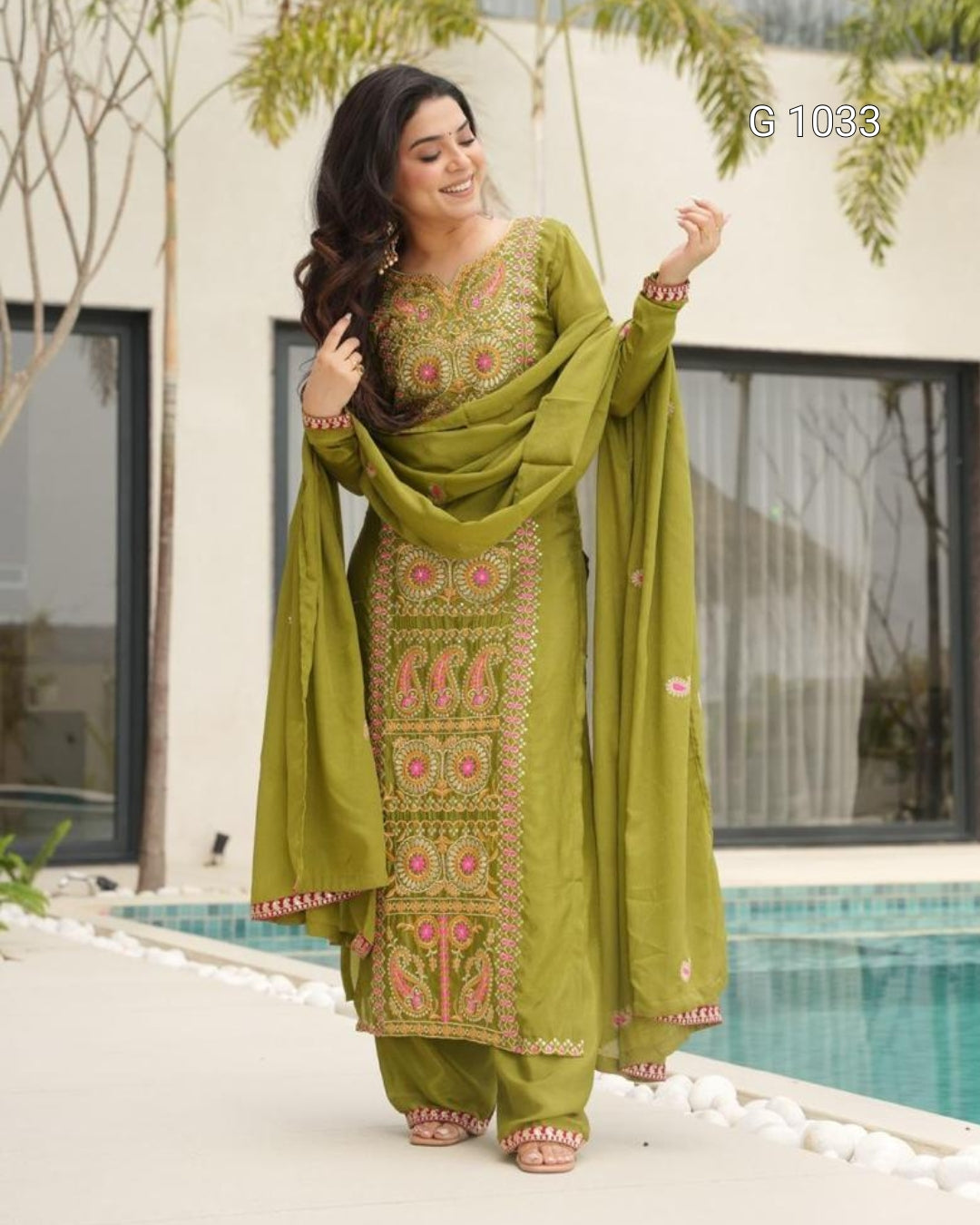 A woman models a traditional Indian salwar kameez suit in olive green with pink and gold embroidery, standing outdoors next to a swimming pool.