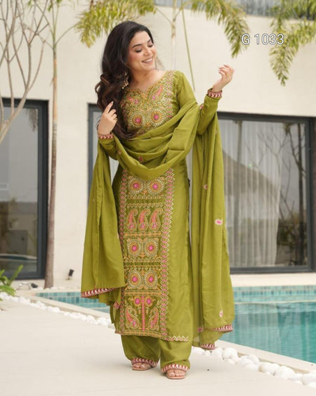 A woman models a traditional Indian salwar kameez suit in olive green with pink and gold embroidery, standing outdoors next to a swimming pool.
