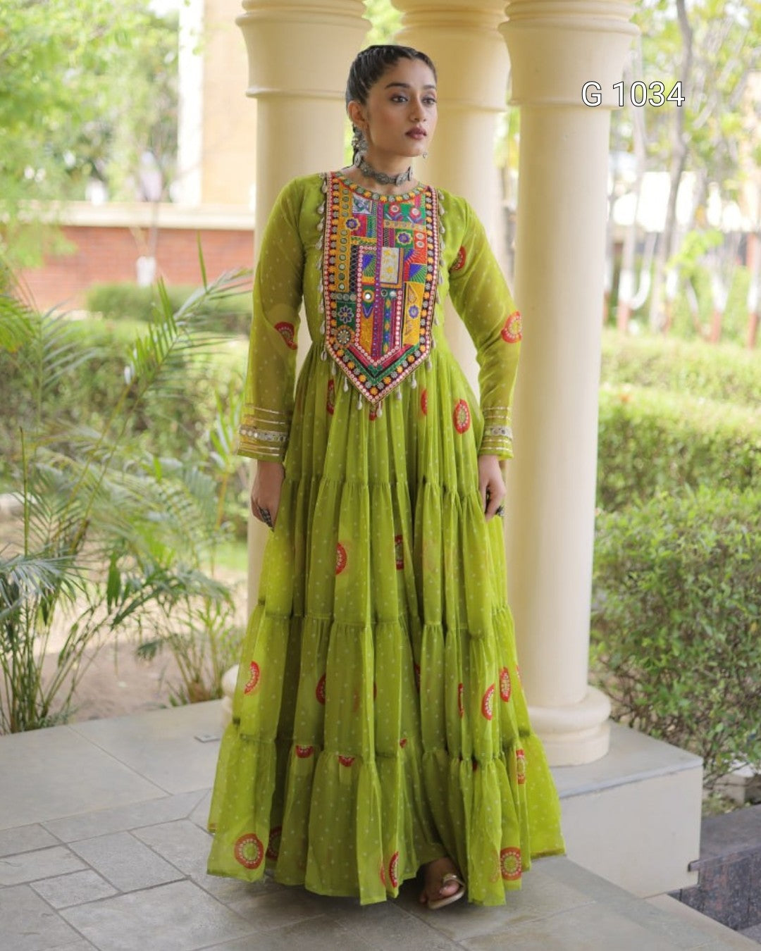 A woman wearing a bright lime green tiered Anarkali gown with colorful embroidered details on the chest and scattered red circular motifs, standing outdoors in front of white pillars and green plants.