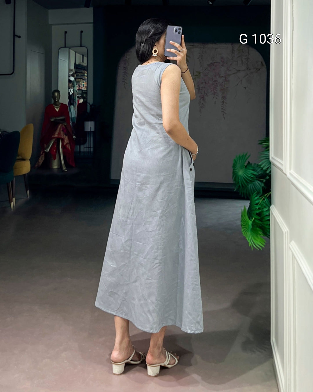 A woman takes a mirror selfie wearing a sleeveless, light gray linen midi dress with a relaxed, flowing silhouette. The dress has a subtle crinkled texture and side pockets.