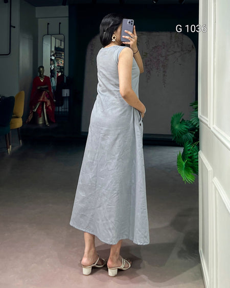 A woman takes a mirror selfie wearing a sleeveless, light gray linen midi dress with a relaxed, flowing silhouette. The dress has a subtle crinkled texture and side pockets.
