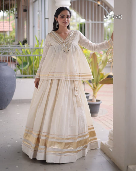 A woman models a traditional Indian outfit consisting of a cream-colored kurta with gold embroidery and a matching flared skirt, set against an outdoor patio background with plants.